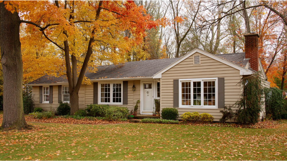 Single-story beige ranch-style home with white trim and a dark shingle roof, surrounded by autumn trees with colorful leaves on the ground.