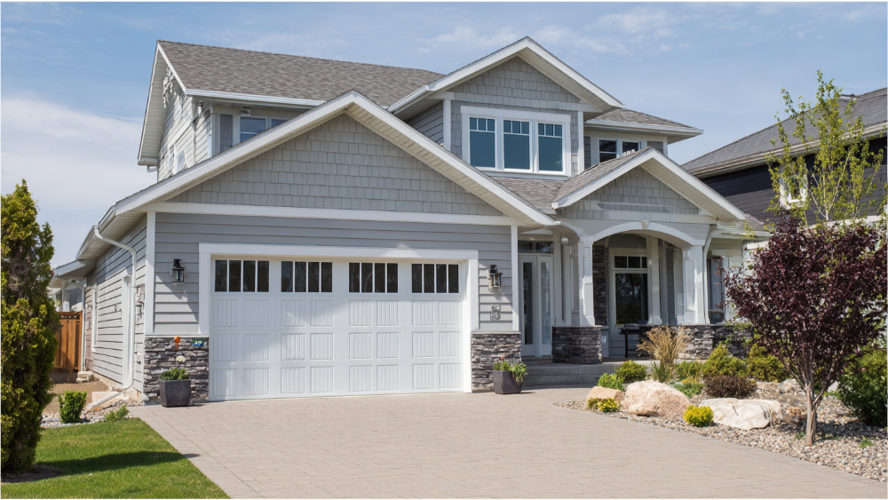 Two-story craftsman-style home with light gray siding, white trim, stone accents, and a wide white garage door, set on a paver driveway under a sunny sky.